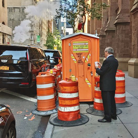 Ein Mann im Anzug vor einer orangefarbenen mobilen Toilette, umgeben von Verkehrspollern in einer belebten Straße mit Dampf aus einer Rohrleitung – Symbolbild für urbane Infrastruktur und Pollerforschung.