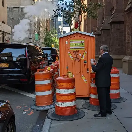 Ein Mann im Anzug vor einer orangefarbenen mobilen Toilette, umgeben von Verkehrspollern in einer belebten Straße mit Dampf aus einer Rohrleitung – Symbolbild für urbane Infrastruktur und Pollerforschung.