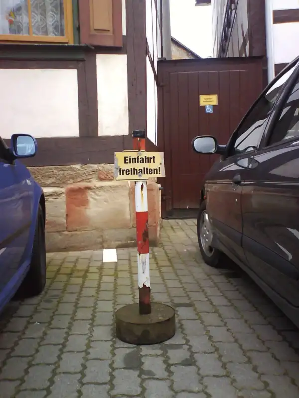 Ein rot-weißer Poller mit einem Schild „Einfahrt freihalten!“ zwischen zwei geparkten Autos in einer engen Hofeinfahrt, typisch für urbane Verkehrsregulierung und Parkplatzmanagement.