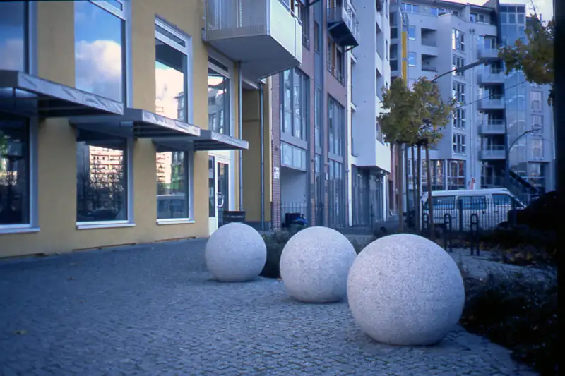 Drei große, runde Poller aus Granit auf einem gepflasterten Bürgersteig vor modernen Wohngebäuden mit großen Glasfenstern, typisches Beispiel für urbane Gestaltung und Verkehrsbarrieren.