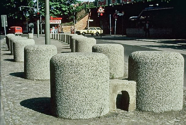 Große, zylinderförmige Betonpoller, die eine Gehwegbegrenzung entlang einer Straße bilden. Im Hintergrund ein Imbiss, parkende Autos und ein Bahndamm. Ein kleiner, gewölbter Durchlass in einem der Poller.