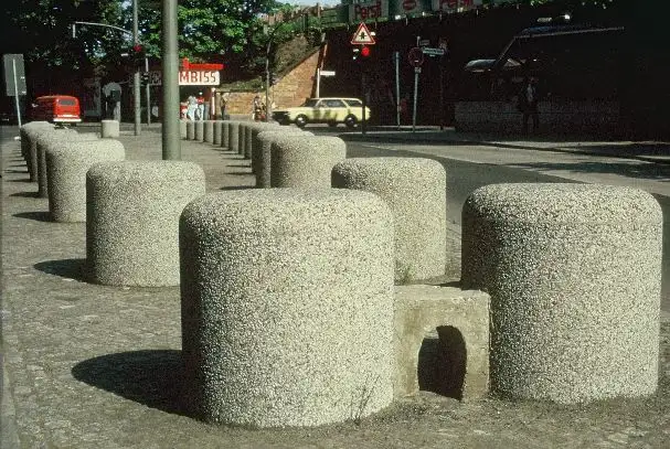 Große, zylinderförmige Betonpoller, die eine Gehwegbegrenzung entlang einer Straße bilden. Im Hintergrund ein Imbiss, parkende Autos und ein Bahndamm. Ein kleiner, gewölbter Durchlass in einem der Poller.