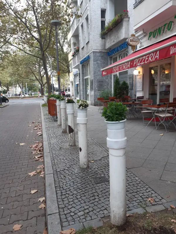 Weiße Verkehrspoller vor einer Trattoria-Pizzeria, dekoriert mit Blumenkübeln, die eine Gehwegbegrenzung entlang eines Baumgesäumten Bürgersteigs bilden.