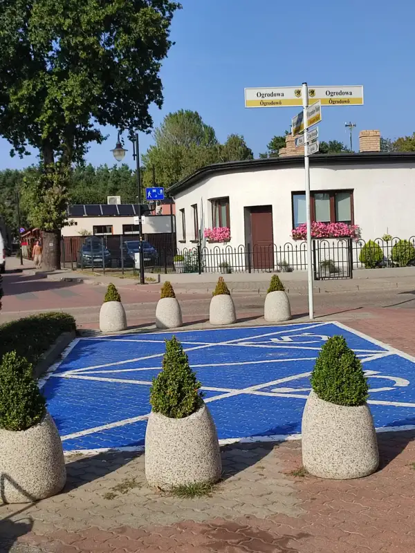 Verkehrspoller in Form von Pflanzkübeln, die einen blau markierten Bereich mit weißem Zebrastreifenmuster in einer ruhigen Wohngegend abgrenzen. Im Hintergrund ein Schild mit Straßennamen und gepflegte Häuser mit Blumenkästen.