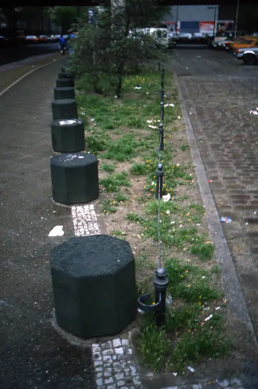 Eine Reihe achteckiger Betonpoller entlang eines Gehwegs, ergänzt durch eine niedrige Metallabsperrung mit Drahtseil, die eine kleine Grünfläche abgrenzt.