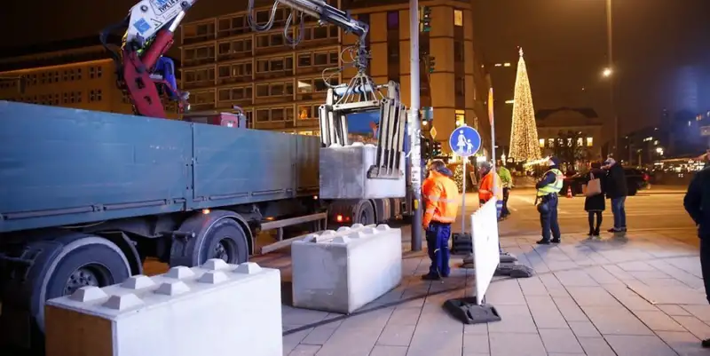 Arbeiter in Sicherheitswesten installieren massive Betonpoller mit Hilfe eines Lastwagens und eines Krans in einer belebten städtischen Umgebung, beleuchtet von Weihnachtslichtern und einem geschmückten Baum im Hintergrund.