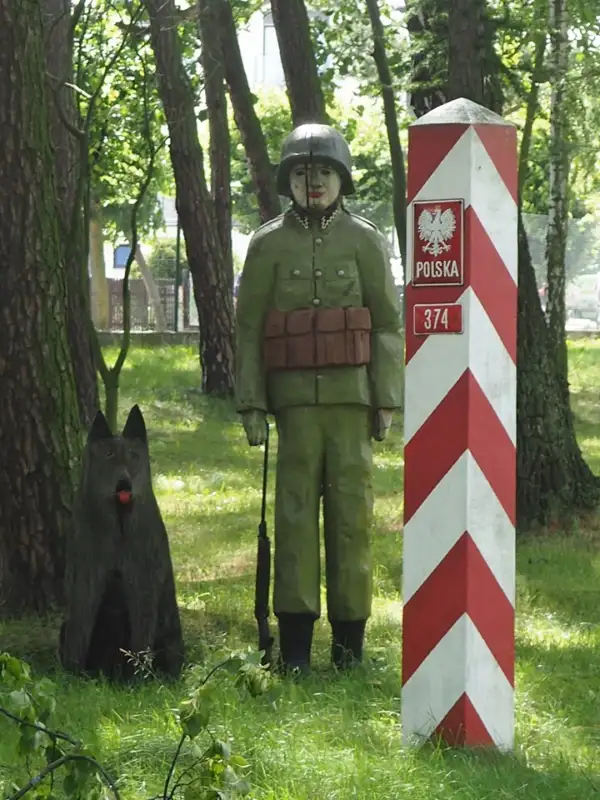 Holzskulptur eines polnischen Grenzsoldaten mit Hund neben einem rot-weißen Grenzpfahl mit der Aufschrift „Polska“ und der Nummer 374. Die Szene befindet sich in einem Waldstück.