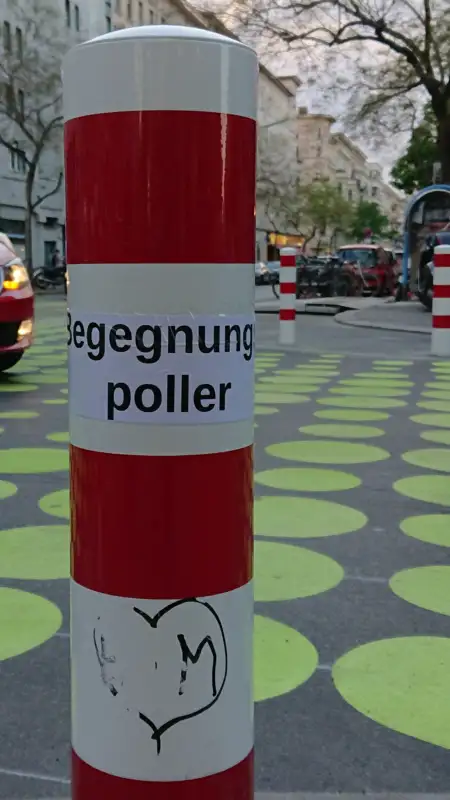 Ein rot-weißer Poller mit der Aufschrift „Begegnung Poller“ steht in einer urbanen Umgebung, umgeben von grünen Kreismustern auf dem Boden – ein Beispiel für Pollerforschung und Stadtgestaltung.