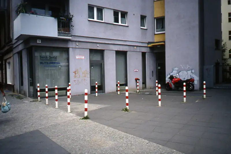 Mehrere rot-weiß gestreifte Poller sichern eine kleine Fläche vor einem Wohn- und Geschäftsgebäude mit Zahnarztpraxis. Im Hintergrund ist ein Motorrad geparkt, und Graffiti zieren die Wände. Dieses Beispiel zeigt die Nutzung von Pollern zur Verkehrs- und Flächensicherung in städtischen Gebieten.